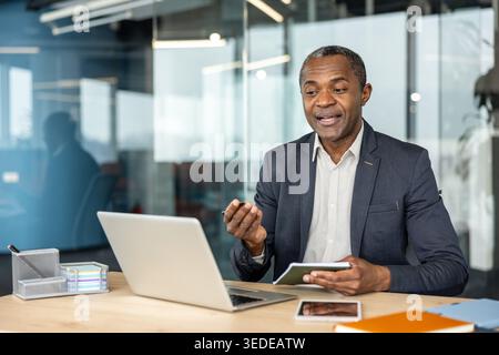 Mature businessman communicating during an online video call at his desk in a modern office, using a laptop and taking notes, representing global communication and remote work Stock Photo