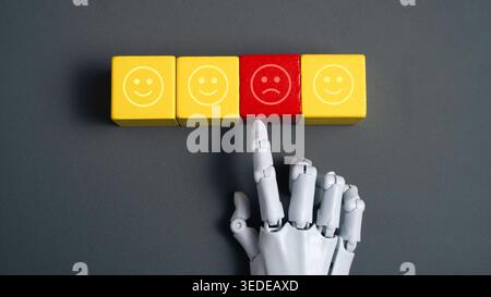 Robot hand selecting a red sad face cube among yellow happy face cubes on a grey background symbolizing AI sentiment analysis and feedback. Stock Photo