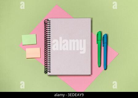 A blank notebook sits open on a table with green and pink paper nearby. Two pens in blue and green lie beside two sticky notes of different colors. Stock Photo