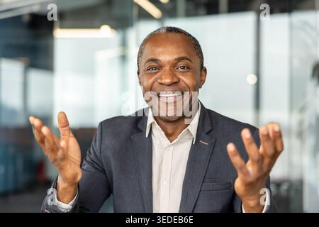 Friendly african american professional man in a suit smiling and gesturing with open hands, conveying confidence, engagement and positive leadership in an office setting Stock Photo