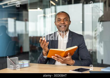 Mature businessman sitting at desk, holding an orange notebook, and making a gesturing motion with a hand, conveying ideas and engaging in thoughtful communication in a corporate setting Stock Photo