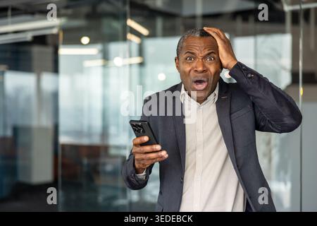 Mature executive looking extremely worried and making a shocked expression with hand on head while receiving, a surprising or stressful message on mobile phone in a modern office Stock Photo