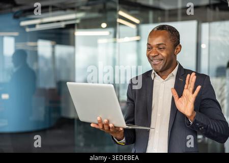 Smiling businessman conducting a virtual meeting or a video conference on his laptop, actively engaging with remote colleagues from a modern office environment Stock Photo