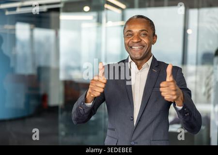 Happy professional man in a suit standing in an office, confidently smiling at the camera and holding both thumbs up, symbolizing approval and success Stock Photo