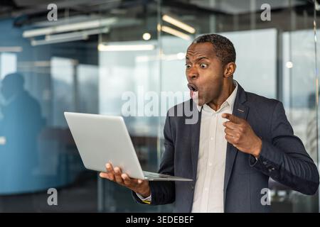 Professional african american man reacting with shock and surprise while pointing at a laptop screen in a modern office, experiencing unexpected success or a sudden revelation Stock Photo