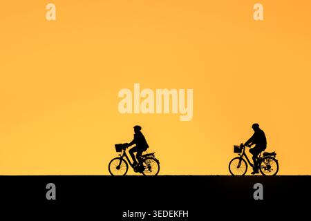 Cycling elderly couple wearing bicycle helmets, riding on their e-bikes / electric bicycles, silhouetted against orange sunset sky in summer Stock Photo