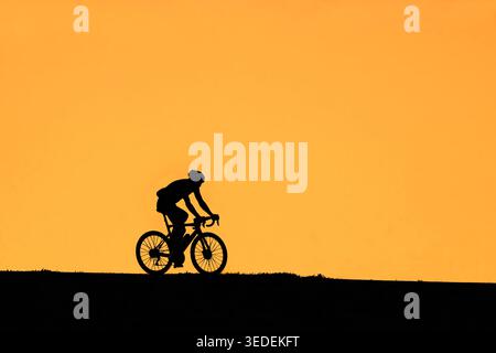 Solitary cyclist riding on his racing bicycle / road bike silhouetted against orange sunset sky in summer Stock Photo