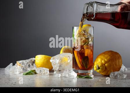 Iced black tea is poured from a bottle into a tall glass with lemon wedges, ice cubes and mint leaves. Gray gradient background with copy space. Stock Photo