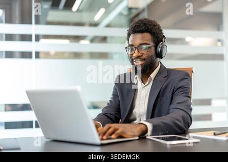 Professional man in glasses and headphones smiling and typing on a laptop in a modern office, focused on remote work or a virtual meeting, conveying productivity and tech confidence Stock Photo