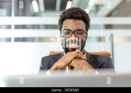 African american businessman wearing optical glasses smiling confidently while using a laptop during work, creating a positive and professional atmosphere in a corporate office setting Stock Photo