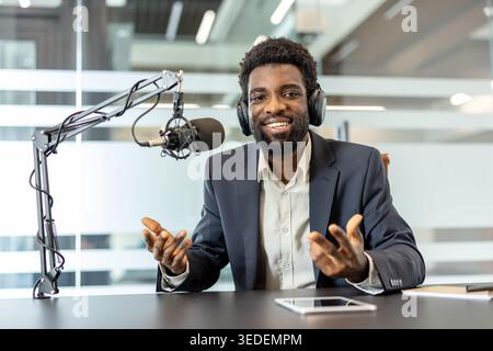 Young man wearing headphones and dark suit, sitting at a desk with a professional microphone, actively gesturing while broadcasting or interviewing in a modern studio environment Stock Photo