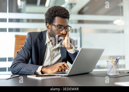 Young businessman wearing glasses and a suit, sitting at a desk with his hand supporting his chin, deeply concentrating on his laptop screen in a modern corporate office setting Stock Photo