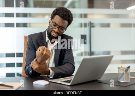 Business man sitting at a desk and holding his painful wrist, suffering from an injury related to repetitive strain and long hours working on a laptop in a modern office environment Stock Photo