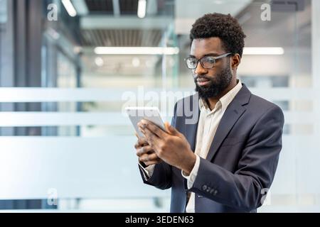 Young professional man with beard and glasses studying data on a digital tablet, focused and confident while managing business tasks in a modern corporate office setting Stock Photo