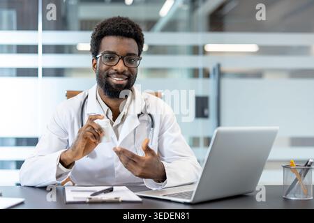Doctor in a white coat with a stethoscope holding a prescription bottle and explaining medication during a virtual healthcare appointment, sitting at a desk with a laptop Stock Photo