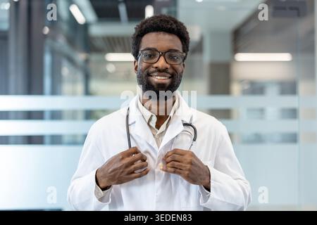 Confident male doctor with afro and beard smiling at camera in white lab coat and stethoscope, standing in a modern clinic conveying friendly, professional healthcare practice Stock Photo