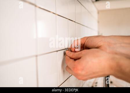 Worker hands carefully placing plastic spacers between white ceramic tiles on a kitchen wall, ensuring even grout lines during a home renovation and construction process Stock Photo