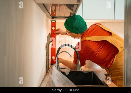 Tiler with a green beanie and mustard overalls precisely installing ceramic tiles on a kitchen backsplash above a sink, using a spirit level for accurate placement during renovation Stock Photo