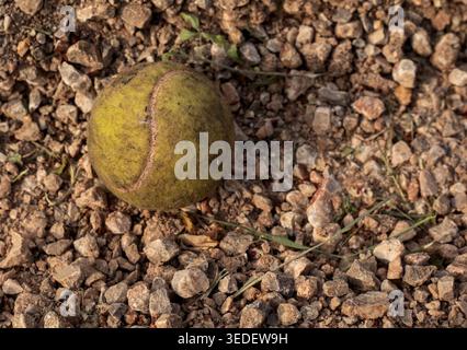 Old dirty yellow tennis ball lying on a pebble and gravel ground. Close-up shot with natural earth tones and rough stone texture. Stock Photo