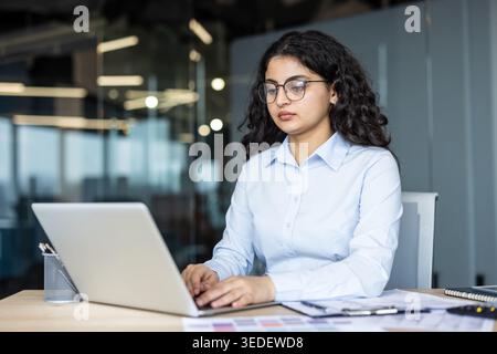 Young indian businesswoman wearing glasses and a blue shirt, concentrating while typing on a laptop computer at a desk in a modern corporate office environment Stock Photo