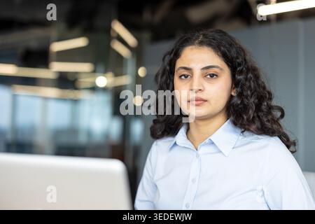 Young professional woman sitting in a modern office, wearing a light blue shirt and looking directly at the camera, representing determination, focus, and corporate success Stock Photo
