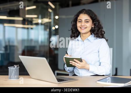 Smiling indian businesswoman sitting at desk in a modern corporate office, holding a notebook and pen while looking at the camera, with a laptop and organized workspace Stock Photo