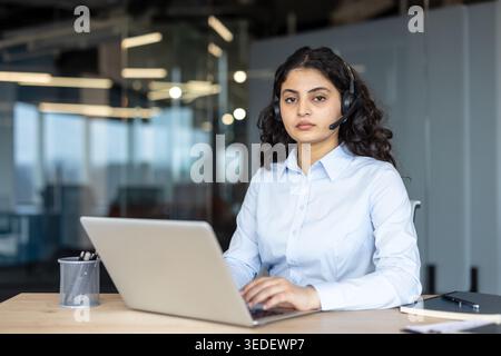 Young female call center operator wearing a professional headset and light blue shirt, working at a desk on a laptop inside a modern office, offering help and communication services Stock Photo