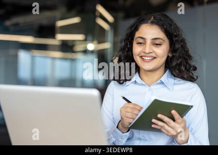 Young indian corporate professional or student smiling while focusing on laptop screen and writing information in a notebook, symbolizing education, training, and remote productivity Stock Photo