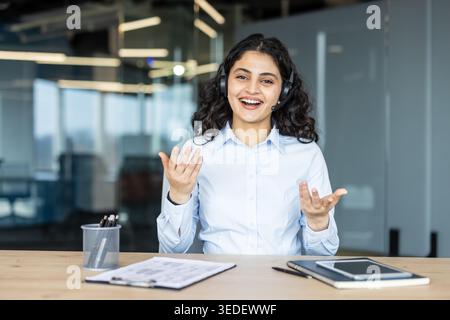 Young woman working as a call center agent or customer support representative, wearing a headset and smiling while talking or presenting in a modern office setting Stock Photo