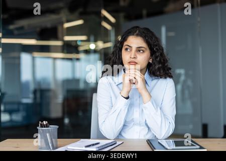 Young professional woman seated at her office desk, gazing away in a thoughtful, focused pose while planning strategies and solutions for career and business growth Stock Photo