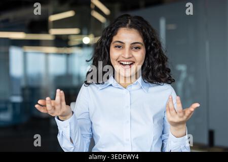 Young indian business woman smiling broadly with open mouth and gesturing with palms up, discussing ideas and sharing joyful communication during a meeting in modern office space Stock Photo