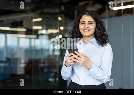 Young smiling businesswoman confidently holding a smartphone, actively engaging with technology, and standing in a modern corporate office environment Stock Photo