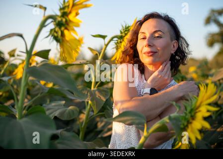 Portrait of a woman in a sunflower field at golden hour, embracing herself and smiling. Warm sunlight and summer vibes convey natural beauty, wellness Stock Photo