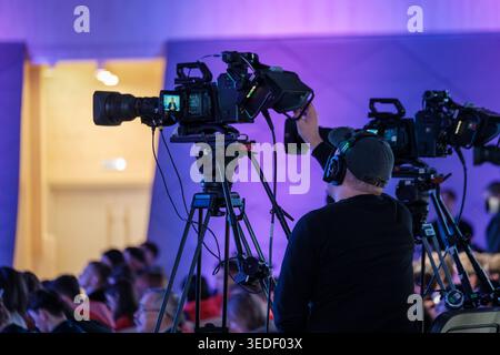 Cameraman operates professional video camera on tripod during live conference, colorful stage lighting and audience. Stock Photo