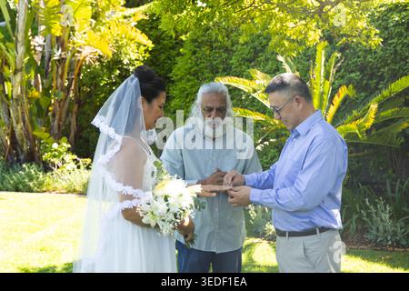 Couple with Indian officiant exchanging rings on sunlit lawn, bride holding white bouquet and veil Stock Photo