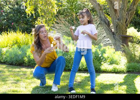 Mother and daughter kneeling blowing soap bubbles with bubble bottles and wands on sunlit lawn Stock Photo