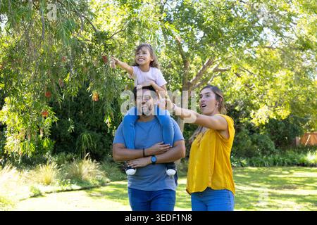 Family trio standing in sunlit park, man carrying child, woman pointing at red flowers Stock Photo