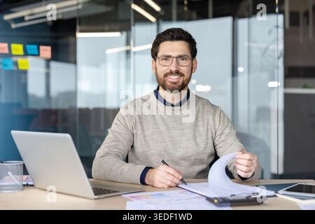 Smiling bearded man with glasses at office desk, holding a pen while reviewing paperwork beside a laptop, confident professional planning and analyzing financial documents Stock Photo