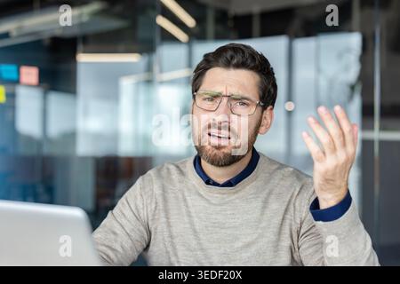 Confused businessman working on a laptop, raising hand, expressing frustration, misunderstanding, and stress, with a displeased facial expression in a modern office environment Stock Photo