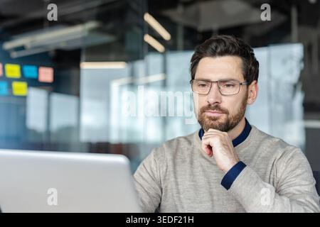 Professional man wearing eyeglasses in a beige sweater and blue shirt concentrating on his work, looking at a modern laptop in a contemporary office setting with glass walls Stock Photo
