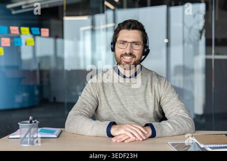 Smiling man in glasses and a headset sitting at an office desk, looking friendly and ready for a video call or providing customer support and online consultation Stock Photo