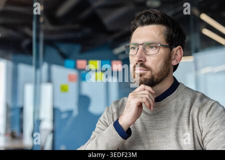 Young male professional in optical glasses pondering a business strategy while working in a modern office, planning solutions with thoughtful concentration Stock Photo