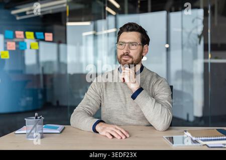 Confident bearded businessman wearing eyeglasses sitting at desk, deep in thought and planning future strategy while working in a contemporary office environment Stock Photo