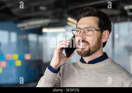 Confident bearded businessman with glasses smiling while talking on his smartphone in a modern office, communicating professionally and staying connected during work Stock Photo
