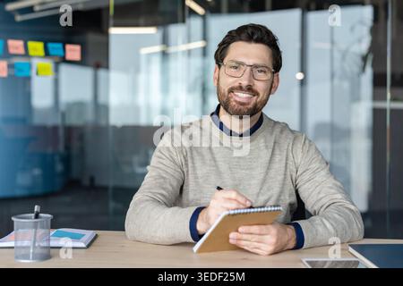 Man with glasses and beard smiling at the camera while sitting at a desk, writing in a notepad, reflecting a positive and professional atmosphere in a contemporary workspace Stock Photo