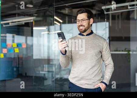 Smiling man wearing glasses and a sweater checking a message on his smartphone, representing communication, technology, and connection in a casual business environment Stock Photo