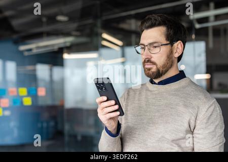 Businessman intently reading information on his smartphone, representing communication, technology, and focusing on digital tasks while working in a modern office environment Stock Photo