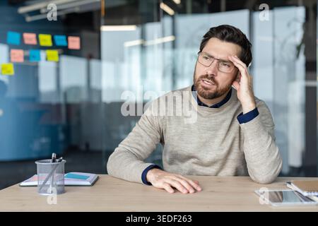 Worried professional man experiencing stress and a painful headache while sitting at an office desk, feeling overwhelmed by problems and work pressure Stock Photo