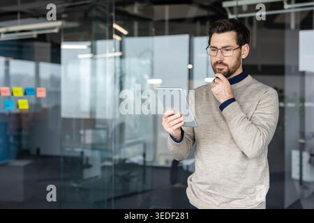 Thoughtful businessman holding a digital tablet in an modern office, contemplating ideas and analyzing information for business strategy and problem-solving, with a hand on his chin Stock Photo