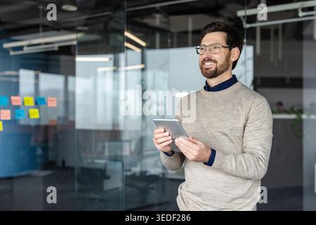 Happy businessman holding a digital tablet and smiling confidently in a modern glass office, envisioning successful future projects and new opportunities Stock Photo
